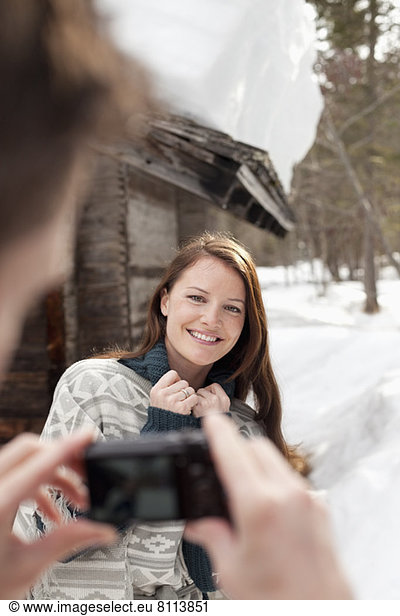 Man photographing smiling woman in snow outside cabin