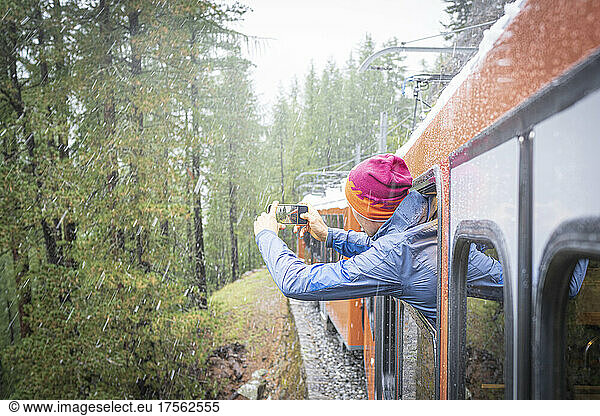 Man photographing the snow falling over the forest leaning out of Gornergrat Bahn train  Zermatt  Valais canton  Switzerland  Europe