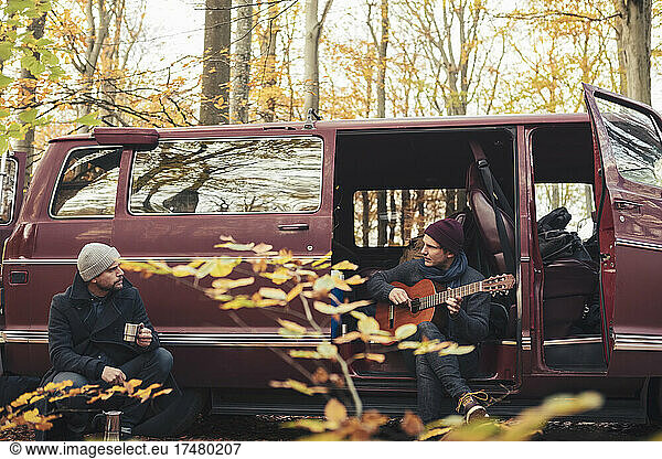 Man playing guitar while male friend having drink in forest during camping