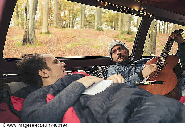 Man plucking guitar while lying by male friend in camping van