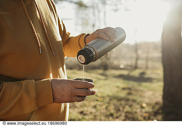 Man pouring tea from insulated drink container in forest on sunny day