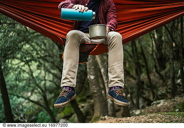 Man pours hot beverage on a pot sitting on a hammock in forest