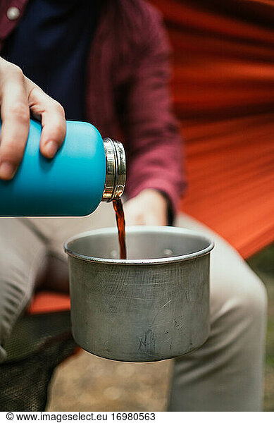 Man pours hot beverage on a pot sitting on a hammock in forest