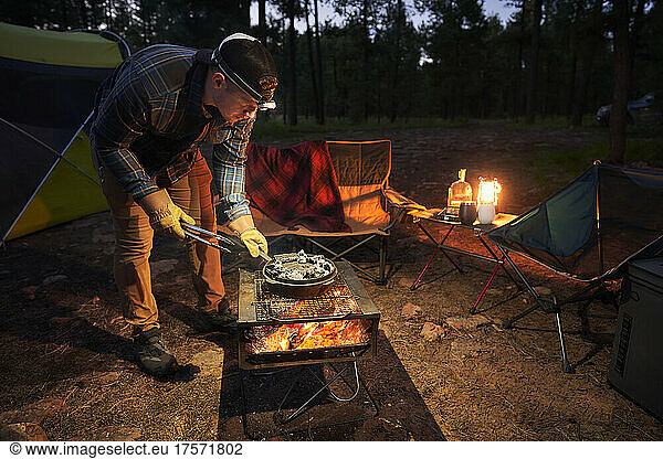 Man prepares camp dinner in a cast iron pan over a fire.