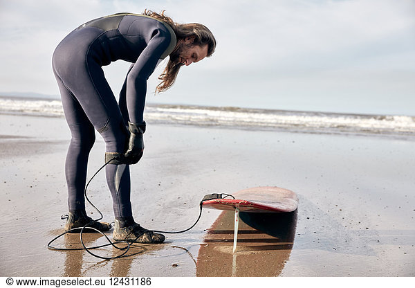 Man preparing to surf on a sandy beach in a wetsuit.