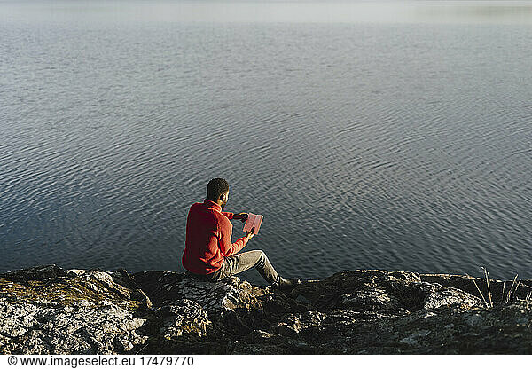 Man reading book while sitting on rock at lakeshore