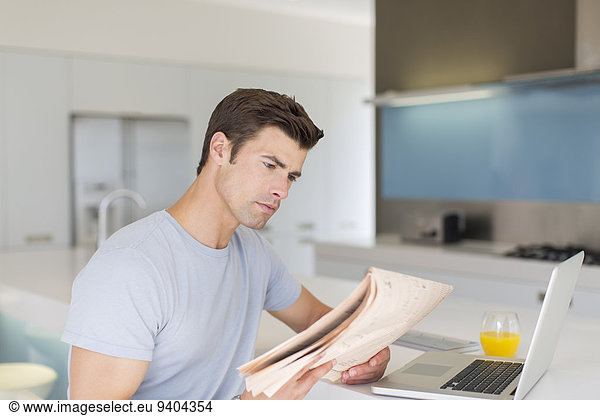 Man reading newspaper in modern kitchen  laptop and orange juice on counter
