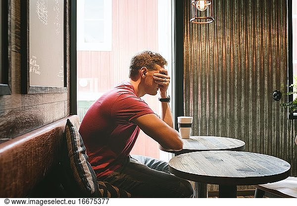 man sat covering his face feeling stressed whilst sat in a cafe