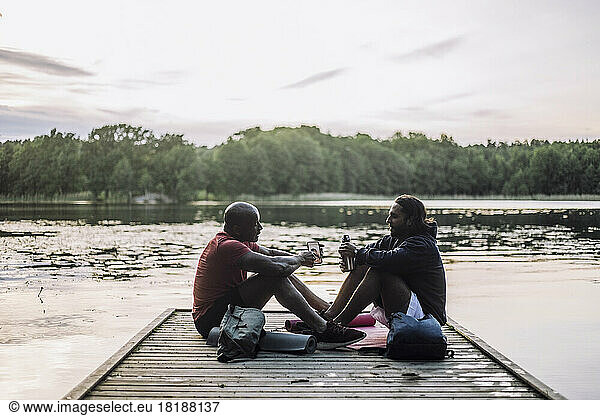 Man sharing smart phone with friend sitting on jetty at lake