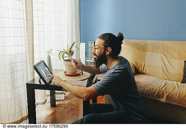 Man showing scissors on video call through tablet PC sitting by sofa at home