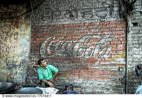 Man sits and looks up at a faded Cola sign painted on a brick wall; Amritsar  Punjab  India