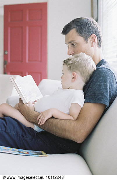 Man sitting on a sofa with his son sitting on his lap  holding a book  reading a story.