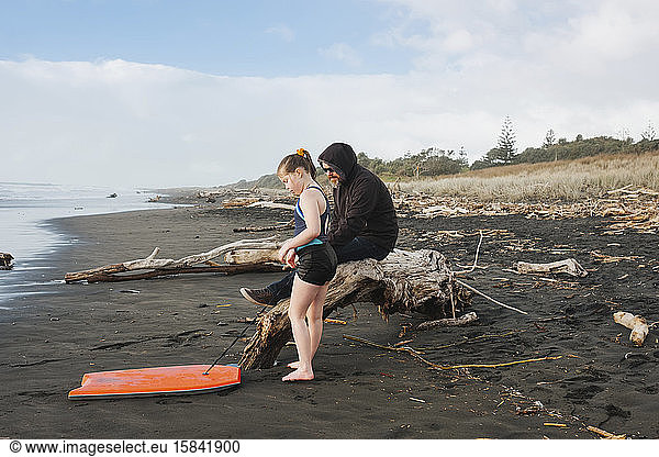 Man sitting on driftwood with girl and her boogie board on the beach