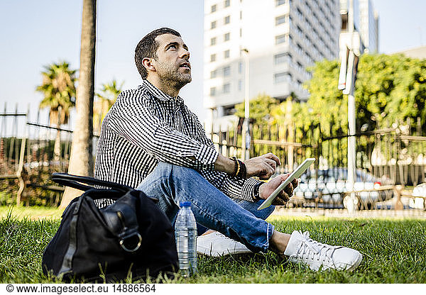 Man sitting on meadow in city park with digital tablet looking up