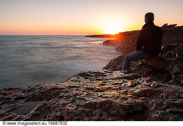 Man sitting on the rocks by the sea watching the sunset. Long exposure
