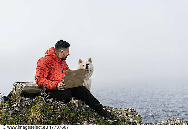 Man sitting with laptop looking at pet dog on rock