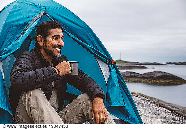 Man smiling holding a mug sitting at a camping tent by the sea