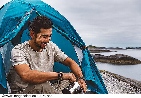 Man smiling pouring coffee in a mug sitting at a camping tent