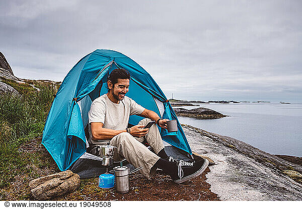 Man smiling typing on a phone while is sitting in a camping tent