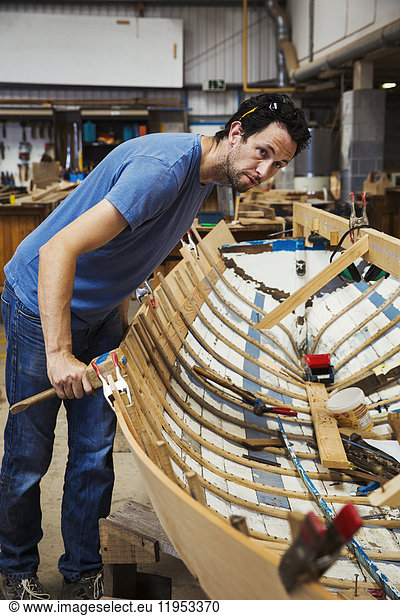 Man standing in a boat-builder's workshop  working on a wooden boat hull.
