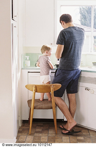 Man standing in a kitchen  his son standing on a chair beside him.