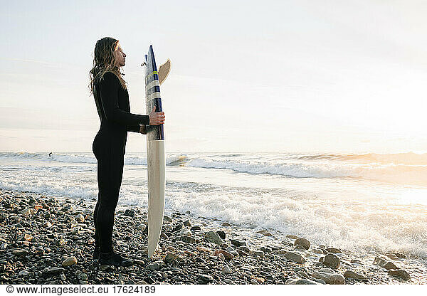 Man standing in wetsuit holding surfboard on rocky beach