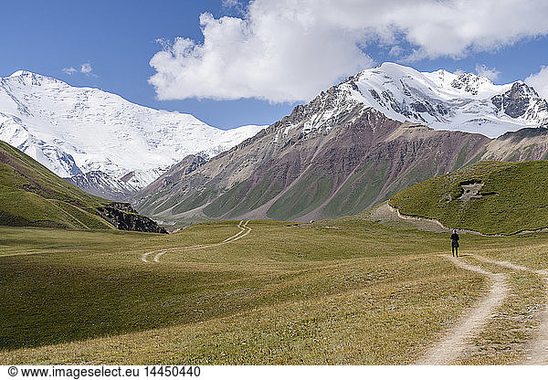 Man standing on tire tracks in a valley surrounded by snow capped mountains  Tulpar Kul  Kyrgyzstan  looking towards Peak Lenin.