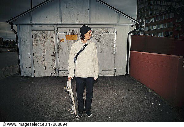 Man standing with skateboard against shed on street