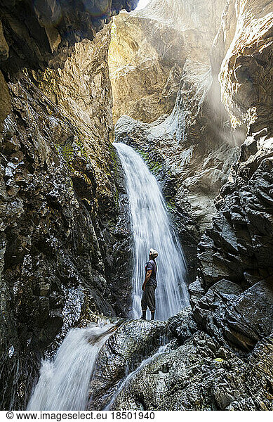 Man stands below waterfall in narrow canyon