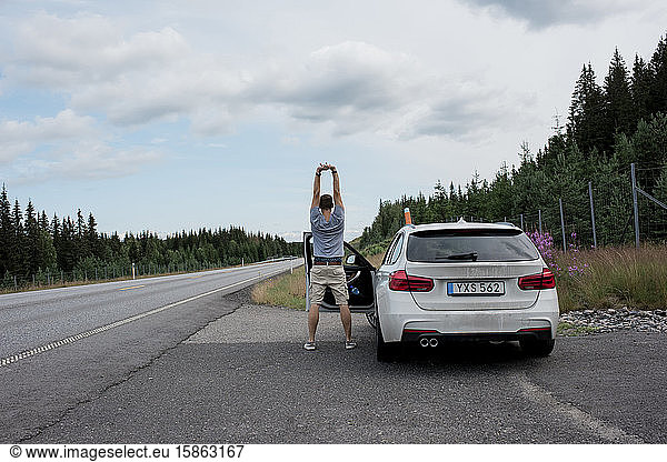 man stretching outside his car by the road after a journey with family
