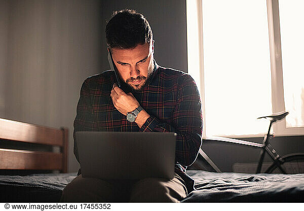 Man talking on smart phone using laptop computer working at home