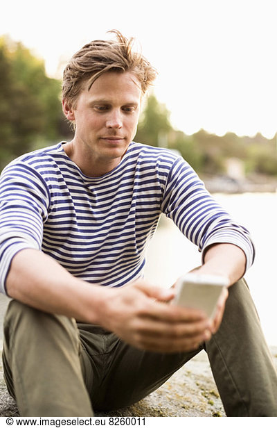 Man text messaging through mobile phone on rock at lake shore