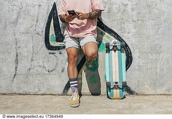 Man text messaging through smart phone by skateboard while leaning on wall during sunny day