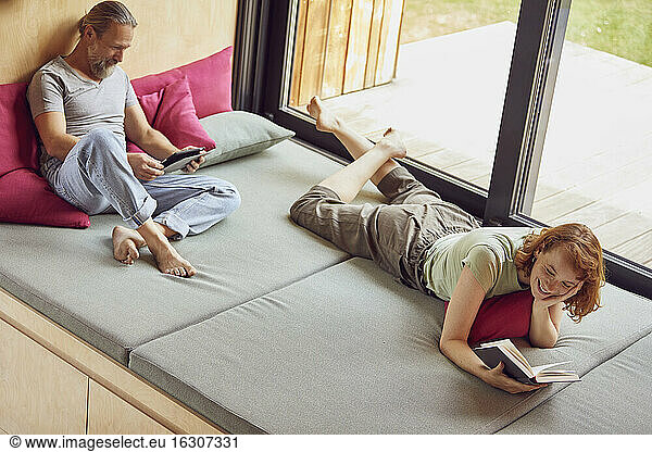 Man using digital tablet while woman reading book on bed at home