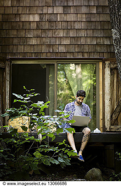 Man using laptop computer while sitting against house