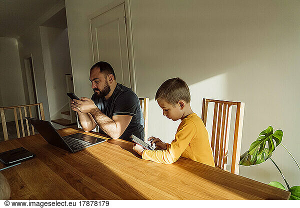 Man using smart phone sitting by son with tablet PC at home