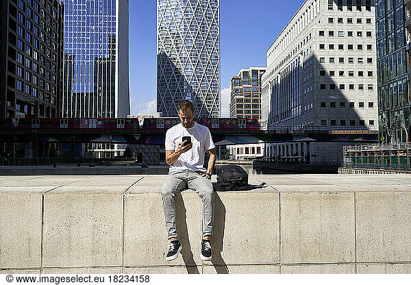 Man using smart phone sitting on wall in front of buildings