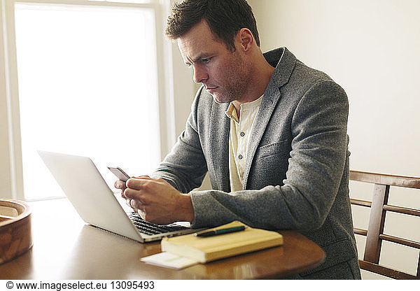 Man using smart phone while sitting with laptop on table at home