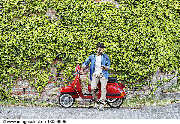Man using smart phone while standing with motor scooter against ivy wall