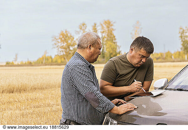 Man using tablet PC on car hood with father in wheat farm