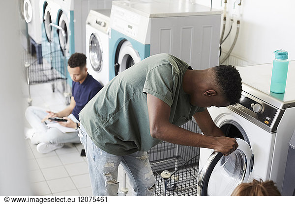 Man using washing machine while university student studying at laundromat