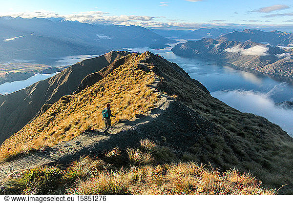 Man walking along ridge line toward peak of mountain overlooking lake.