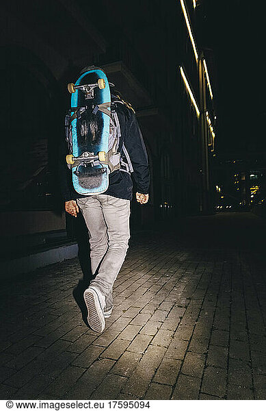Man walking with backpack and skateboard walking on footpath at night