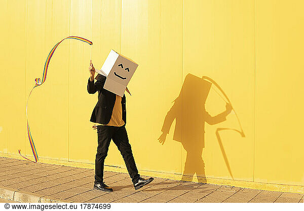 Man wearing box with face spinning rainbow colored rhythmic gymnastics ribbon walking by yellow wall on footpath