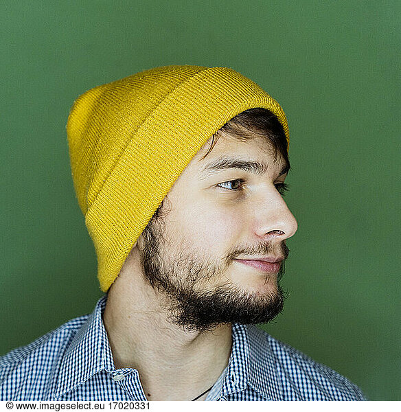 Man wearing knit hat looking away while standing against green background