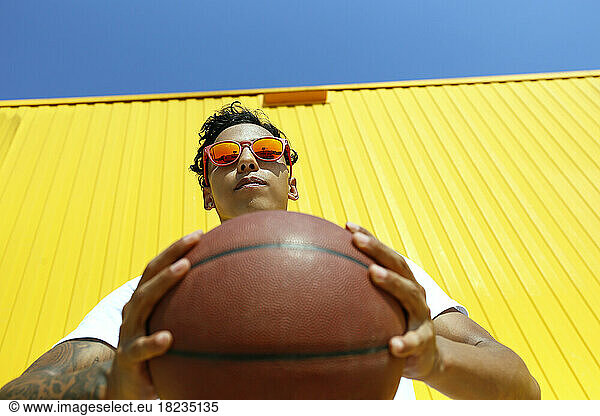 Man wearing red sunglasses holding basketball in front of yellow wall