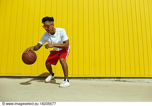 Man wearing red sunglasses playing with basketball on footpath in front of yellow wall