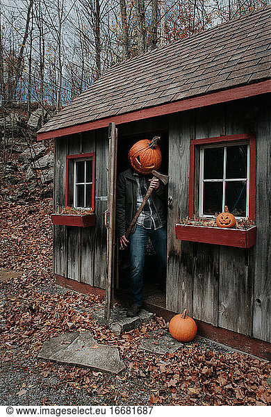 Man wearing scary carved pumpkin head in shed with axe for Halloween.