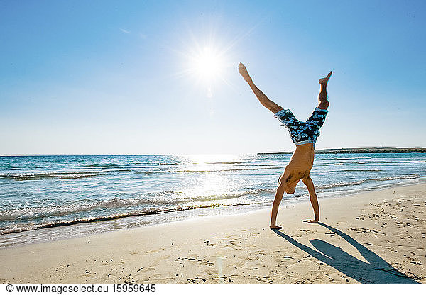 Man wearing swimming shorts doing cartwheel on sandy beach in Mediterranean.