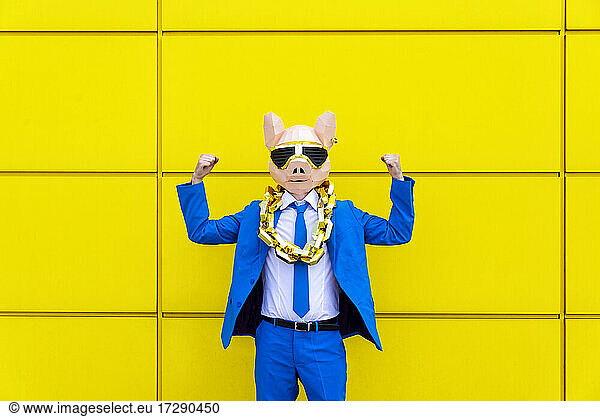 Man wearing vibrant blue suit  pig mask and large golden chain flexing muscles against yellow wall